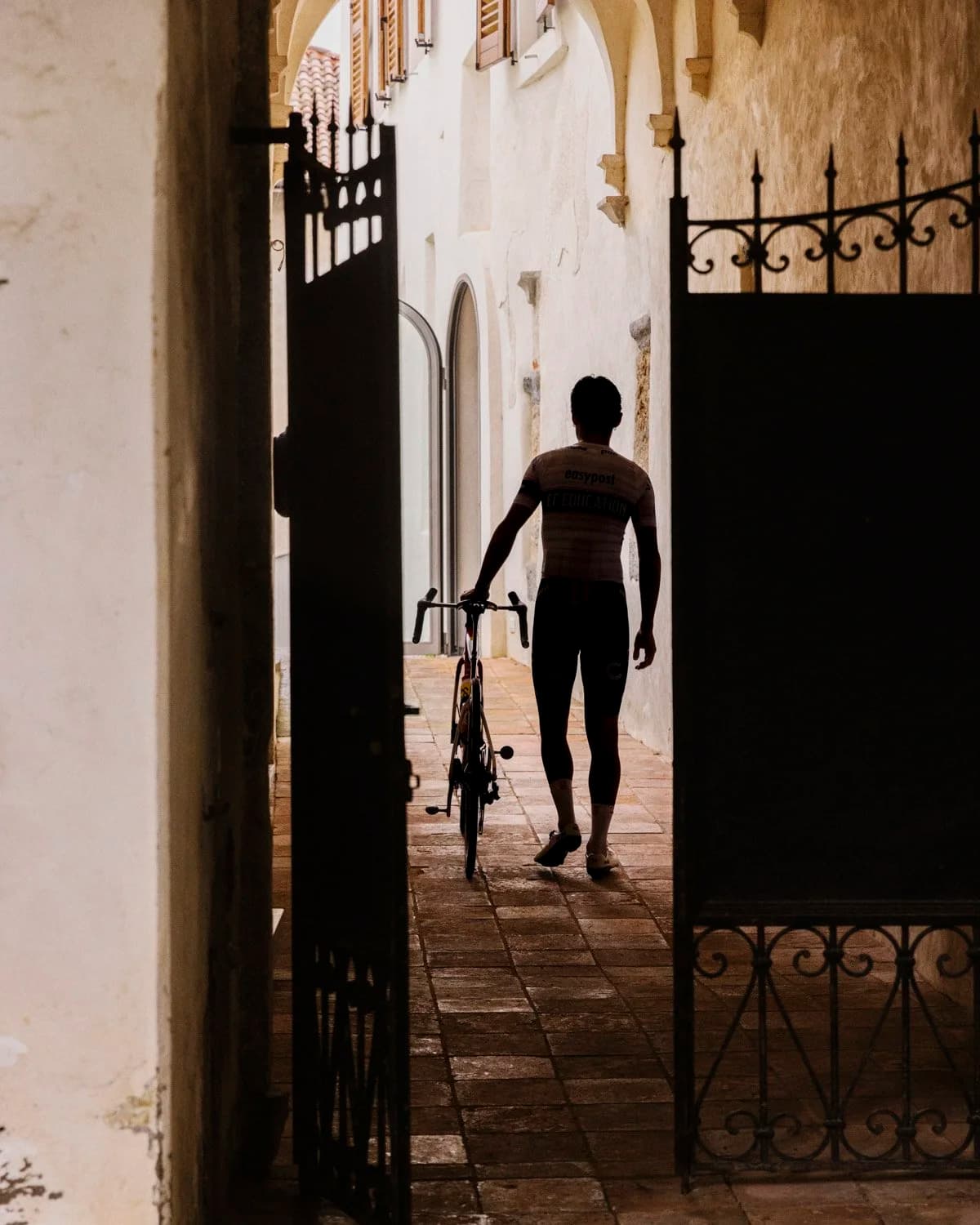 A cyclist silhouetted walking his bike through a sunlit Italian archway, photographed by David Powell